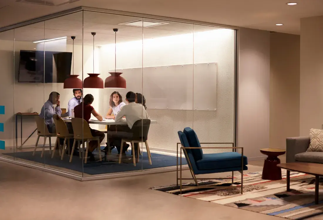 A diverse team engaged in a discussion within a modern glass-walled meeting room, working together collaboratively to better overcome self-limiting beliefs and to build resilience together.