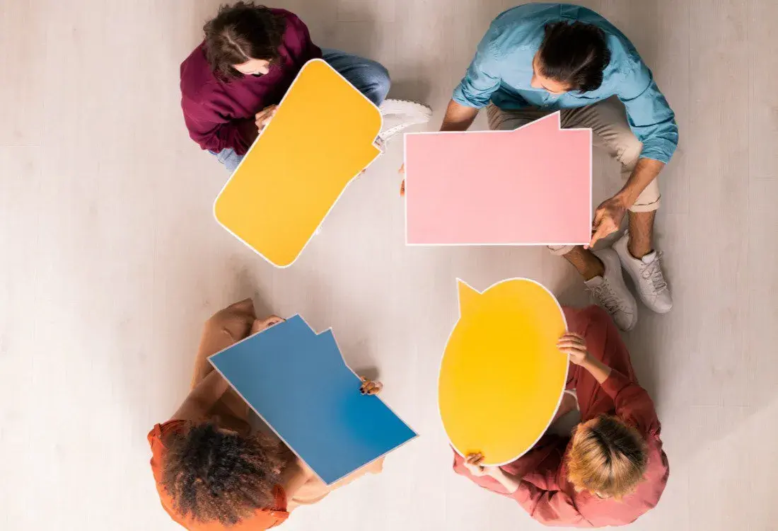 Group of people sitting in a circle holding colourful speech bubbles, symbolising open dialogue, active listening, and the value-building behaviours essential for effective communication.